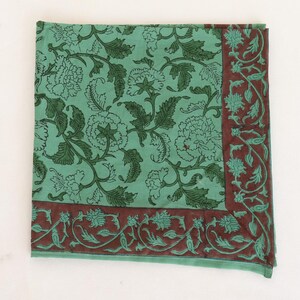 May include: A square, folded textile with a floral pattern. The main color is a light teal, with dark green floral designs. A border of dark red-brown with teal floral accents frames the edges. The textile is likely cotton or linen.