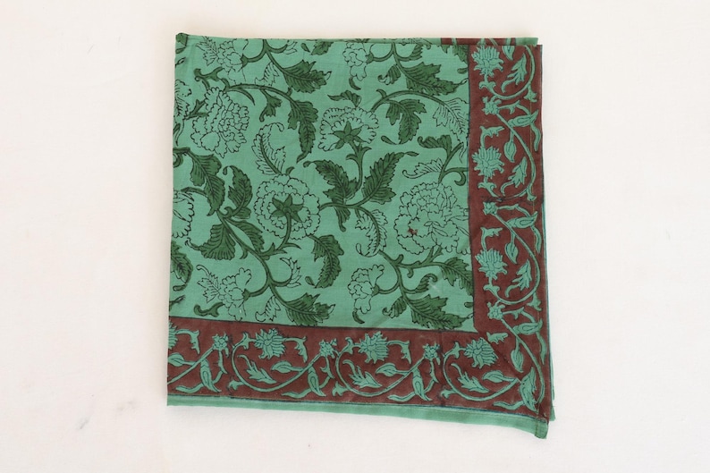 May include: A square, folded textile with a floral pattern. The main color is a light teal, with dark green floral designs. A border of dark red-brown with teal floral accents frames the edges. The textile is likely cotton or linen.