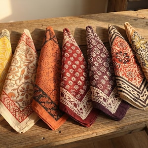 Könnte beinhalten: Eine Sammlung von sieben bunten, gemusterten Stoffservietten, die auf einem rustikalen Holztisch angeordnet sind. Die Servietten weisen verschiedene Blumen- und geometrische Designs in Gelb-, Orange-, Rot- und Lilatönen mit dekorativen Bordüren auf.