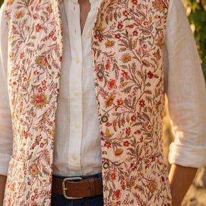 May include: A cream-colored quilted vest with a floral pattern in red, yellow, and green. The vest is buttoned over a white button-down shirt and paired with blue jeans and a brown belt. The vest has a quilted texture.