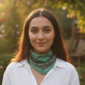 May include: A person wearing a white button-down shirt and a green bandana with a paisley design. The bandana is around the neck. The background is a garden with soft sunlight.