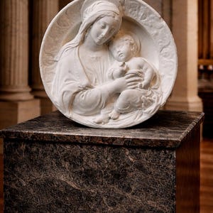 May include: A white marble relief sculpture of the Madonna and Child. The sculpture is circular, with the figures in high relief. The Madonna is depicted with a halo, cradling the child. The sculpture rests on a dark marble-topped pedestal.