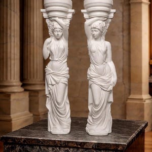 Puede incluir: Dos esculturas blancas de figuras femeninas clásicas, cada una sosteniendo un capitel. Las figuras están envueltas en túnicas fluidas y se alzan sobre una superficie de mármol oscuro. El fondo presenta columnas y detalles arquitectónicos.