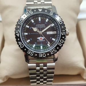 May include: A stainless steel Citizen automatic watch with a black rotating bezel and a dark brown face. The watch features a date display and a silver link band. The watch is displayed on a beige cushion.