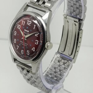 May include: A silver-toned wristwatch with a burgundy dial. The watch features a stainless steel band and a round face with white numerals and hands. The dial includes the text "hmt PILOT" and "17 JEWELS".
