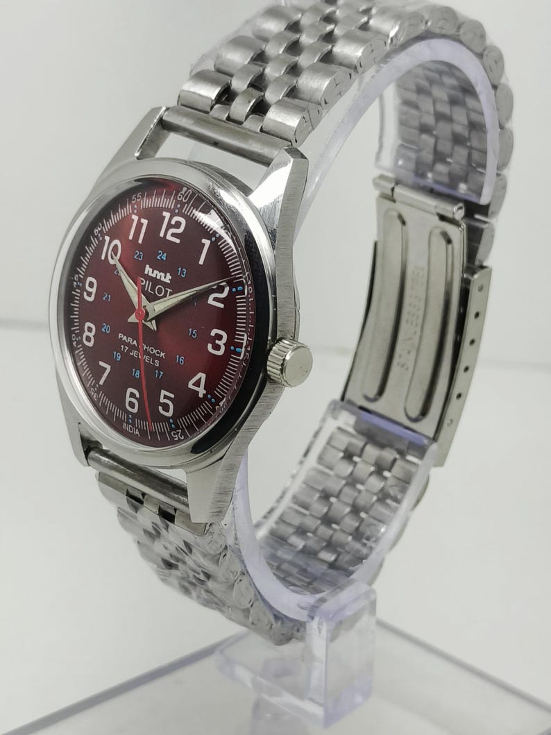 May include: A silver-toned wristwatch with a burgundy dial. The watch features a stainless steel band and a round face with white numerals and hands. The dial includes the text "hmt PILOT" and "17 JEWELS".