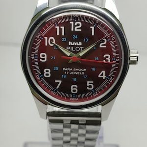 May include: A silver wristwatch with a burgundy dial featuring white numerals and hands. The watch face displays the text "hmt PILOT" and "PARA SHOCK 17 JEWELS". The watch has a silver band.
