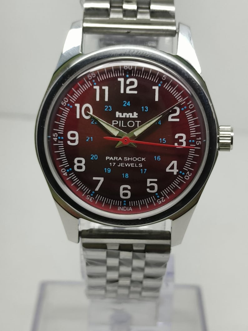 May include: A silver wristwatch with a burgundy dial featuring white numerals and hands. The watch face displays the text "hmt PILOT" and "PARA SHOCK 17 JEWELS". The watch has a silver band.