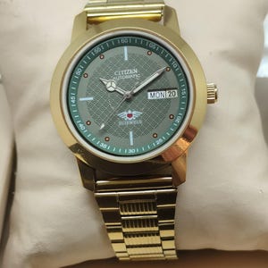 Könnte beinhalten: Goldfarbene Citizen Automatic Uhr mit grünem Zifferblatt und goldenem Armband. Das Zifferblatt zeigt den Tag und das Datum mit dem Text "MON 20". Die Uhr wird auf einem cremefarbenen Kissen präsentiert.