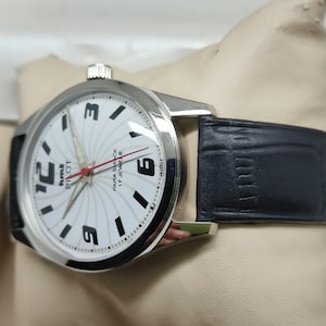 May include: Silver-tone wristwatch with a white face featuring black numerals and hands. The watch has a black leather band. The watch face reads "Pilot Para Shock 17 Jewels".