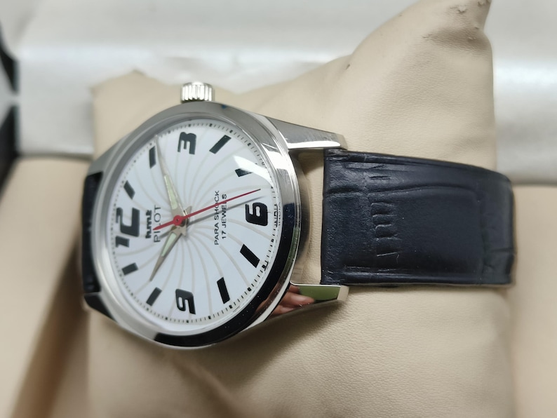 May include: Silver-tone wristwatch with a white face featuring black numerals and hands. The watch has a black leather band. The watch face reads "Pilot Para Shock 17 Jewels".