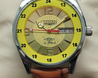 May include: A vintage Citizen automatic watch with a yellow face and a brown leather band. The watch features a gold-toned dial with the day and date displayed. The watch has a silver-colored case and a brown leather strap.