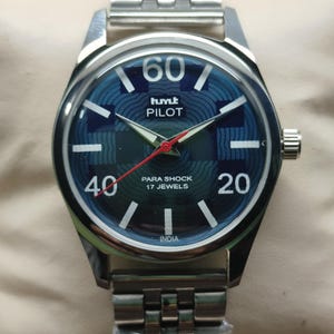 Pode incluir: Um relógio de pulso prateado com mostrador azul estampado. O mostrador exibe os textos "60", "hmt PILOT", "40" e "20". O relógio tem uma pulseira prateada e um ponteiro de segundos vermelho. O texto "PARA SHOCK 17 JEWELS" e "INDIA" também são visíveis.