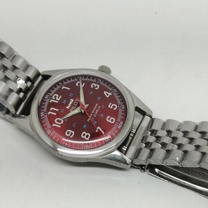 May include: Silver-toned wristwatch with a red dial featuring white numerals and hands. The watch has a stainless steel band and the text "hmk PILOT" is visible on the dial. The watch is new and still has the protective plastic on the band.