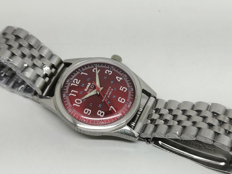 May include: Silver-toned wristwatch with a red dial featuring white numerals and hands. The watch has a stainless steel band and the text "hmk PILOT" is visible on the dial. The watch is new and still has the protective plastic on the band.
