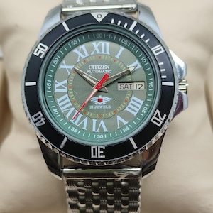 Pode incluir: Um relógio de pulso Citizen Automatic com pulseira prateada e moldura giratória preta. O mostrador é verde com algarismos romanos e ponteiros vermelhos. O dia é exibido como "SAT 12".