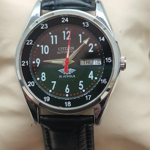May include: A Citizen automatic wristwatch with a black leather band. The watch face is black with white numerals and red accents. The watch displays the day and date. The watch has a silver-colored bezel and case.