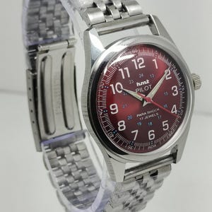 May include: A silver wristwatch with a red dial. The watch face displays white numerals and hands, with the text "hmt PILOT" and "PARA SHOCK 17 JEWELS". The watch has a stainless steel band.