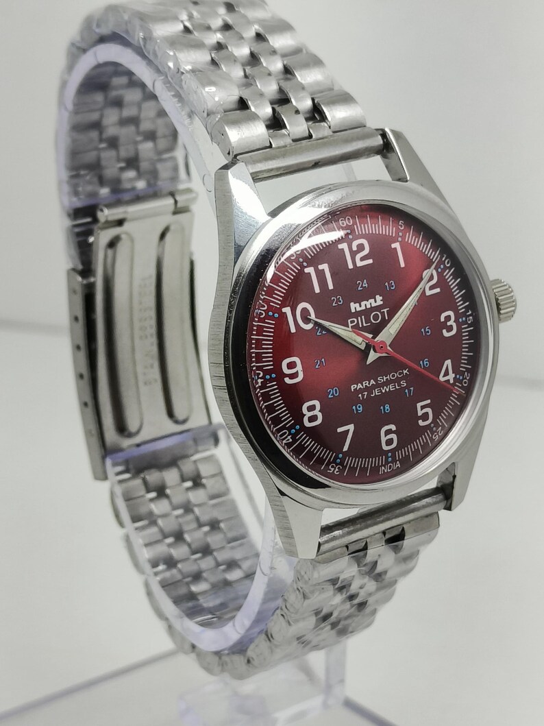 May include: A silver wristwatch with a red dial. The watch face displays white numerals and hands, with the text "hmt PILOT" and "PARA SHOCK 17 JEWELS". The watch has a stainless steel band.