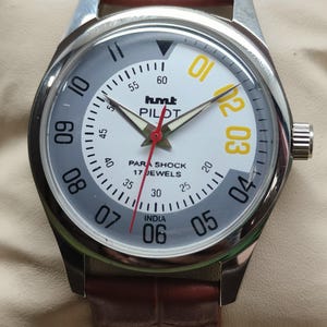 May include: A silver wristwatch with a white and grey face, displaying yellow and white numerals. The watch has a brown leather strap and the text "hmt PILOT" is visible. The watch face also reads "PARA SHOCK 17 JEWELS INDIA".