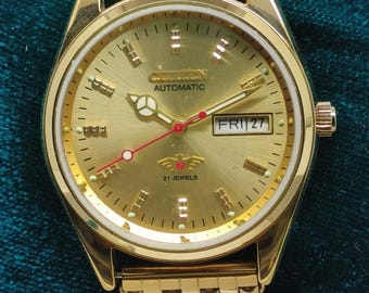 May include: A gold-toned Citizen automatic wristwatch with a matching gold band. The watch face displays the day and date, with the text "Citizen Automatic" and "21 Jewels". The watch has a gold bezel and a red second hand.