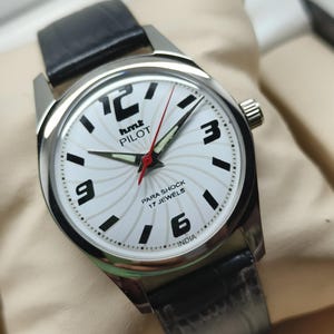 May include: A silver-toned wristwatch with a white face featuring black numerals and hands. The watch has a black leather band and a red second hand. The watch face includes the text "hmt PILOT" and "PARA SHOCK 17 JEWELS INDIA".