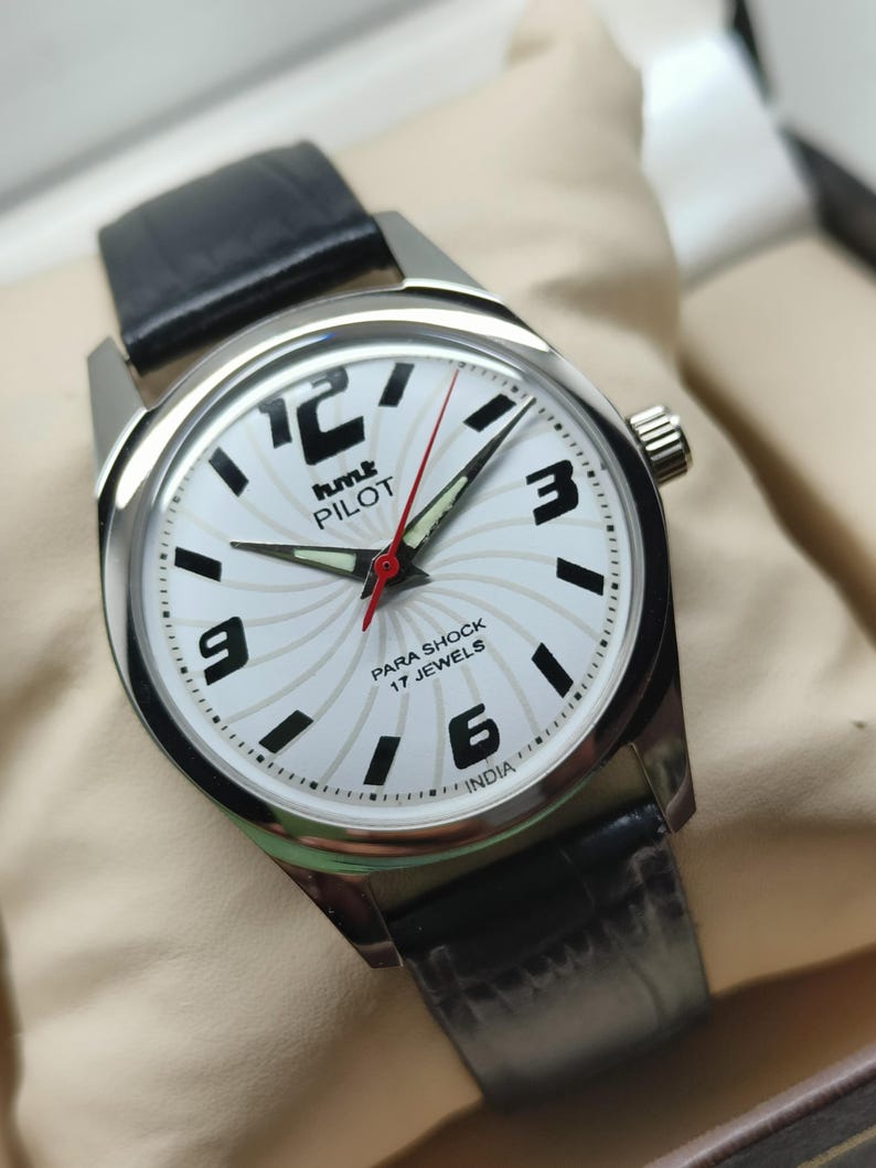 May include: A silver-toned wristwatch with a white face featuring black numerals and hands. The watch has a black leather band and a red second hand. The watch face includes the text "hmt PILOT" and "PARA SHOCK 17 JEWELS INDIA".