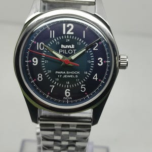 Puede incluir: Un reloj de pulsera plateado con esfera azul oscuro. El reloj tiene una esfera redonda con números blancos, un segundero rojo y el texto "hmt PILOT" y "PARA SHOCK 17 JEWELS INDIA". La correa es de acero inoxidable.