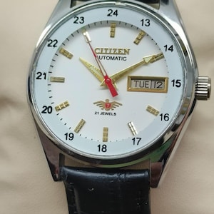 May include: A silver-toned Citizen automatic watch with a white face, black leather band, and gold-colored hands and markers. The watch face displays the day and date, with the text "CITIZEN AUTOMATIC" and "21 JEWELS".