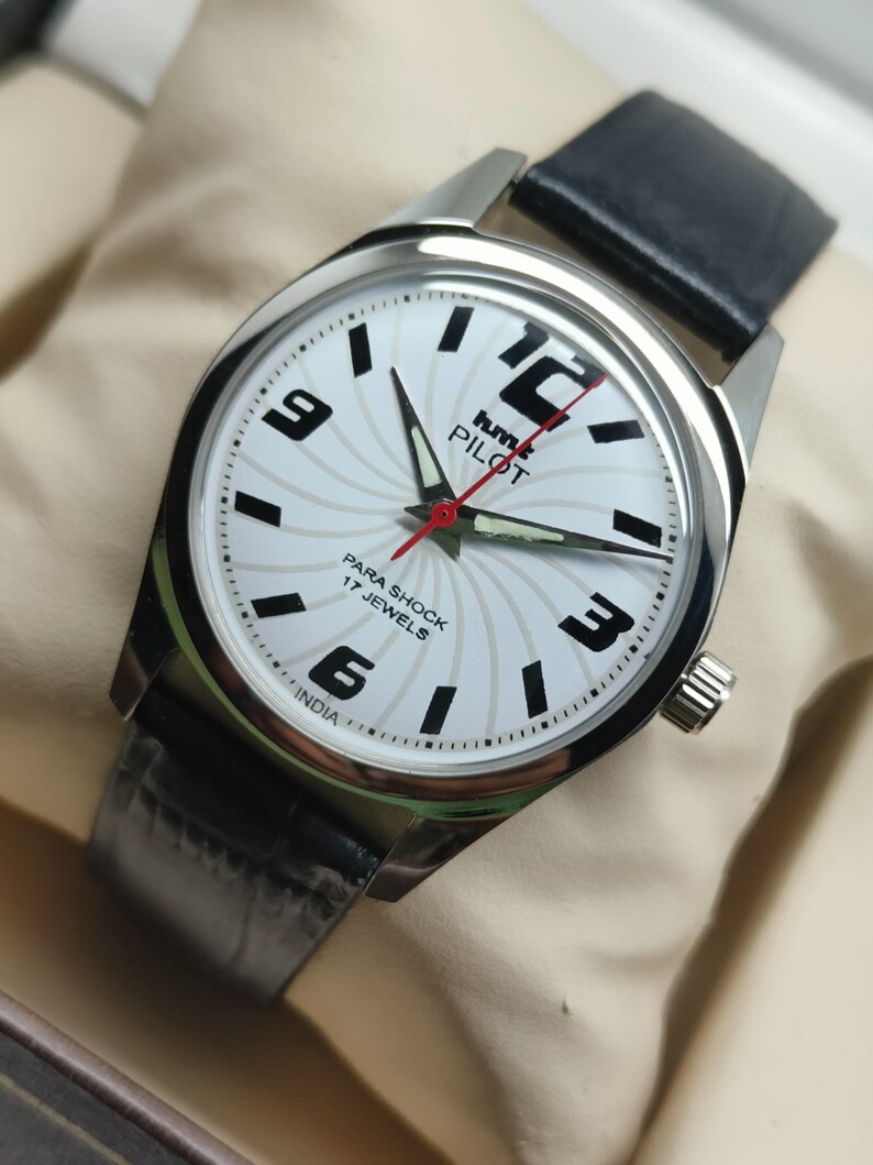 May include: Silver-toned wristwatch with a white face featuring black numerals and hands. The watch has a black leather band and a red second hand. The watch face has the text "hmt PILOT" and "PARA SHOCK 17 JEWELS INDIA".