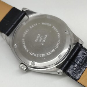 May include: Close-up of the back of a wristwatch with a stainless steel case and a black leather strap. The watch face is silver with text that reads "STAINLESS STEEL BACK - WATER DUST PROTECTED - SHOCK RESISTANT".
