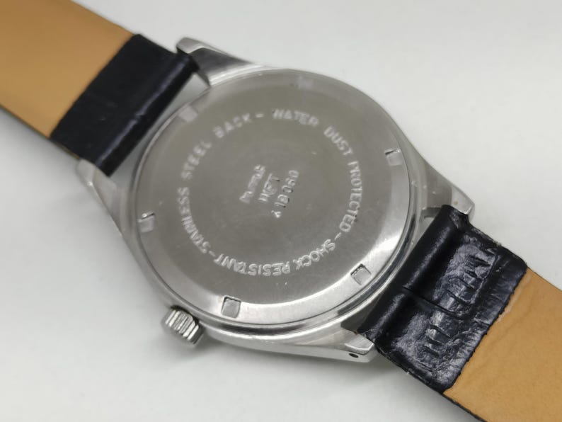 May include: Close-up of the back of a wristwatch with a stainless steel case and a black leather strap. The watch face is silver with text that reads "STAINLESS STEEL BACK - WATER DUST PROTECTED - SHOCK RESISTANT".