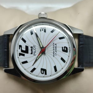 May include: A silver-toned wristwatch with a white face featuring black numerals and hands. The watch has a black leather band and the text "hmt PILOT" and "PARA SHOCK 17 JEWELS INDIA" are visible on the watch face.