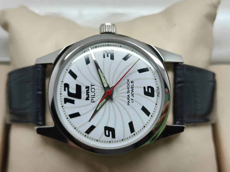 May include: A silver-toned wristwatch with a white face featuring black numerals and hands. The watch has a black leather band and the text "hmt PILOT" and "PARA SHOCK 17 JEWELS INDIA" are visible on the watch face.