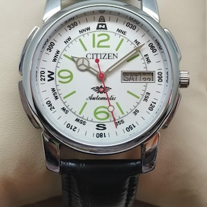 Puede incluir: Un reloj de pulsera automático Citizen con esfera blanca, correa de cuero negro y caja plateada. La esfera del reloj presenta números verdes, una pantalla de día y un bisel estilo brújula. Las manecillas del reloj son rojas y plateadas.