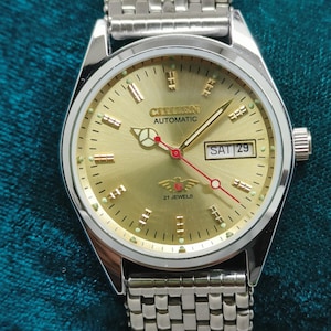 Pode incluir: Um relógio Citizen Automatic dourado com pulseira prateada. O mostrador mostra o dia e a data, com o texto "Citizen Automatic" e "21 Jewels". O relógio tem um ponteiro de segundos vermelho e está definido para sábado, dia 29.