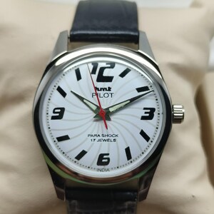 May include: A silver-toned wristwatch with a white face featuring black numerals and hands. The watch face has the text "hmt PILOT PARA SHOCK 17 JEWELS INDIA". The watch has a black leather band.