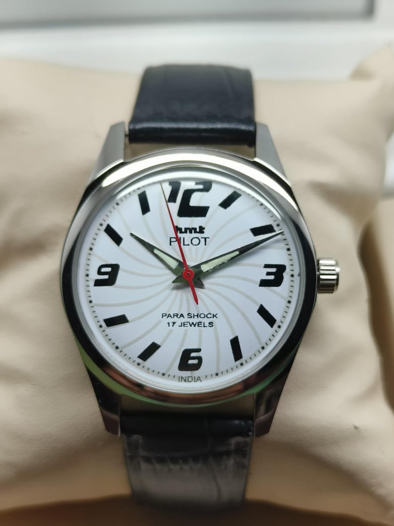May include: A silver-toned wristwatch with a white face featuring black numerals and hands. The watch face has the text "hmt PILOT PARA SHOCK 17 JEWELS INDIA". The watch has a black leather band.