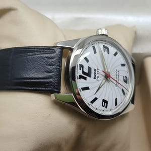 May include: A silver-toned wristwatch with a black leather band. The watch face is white with black numerals and hands, and the text "Pilot" is visible. The watch is displayed on a cream-colored surface.