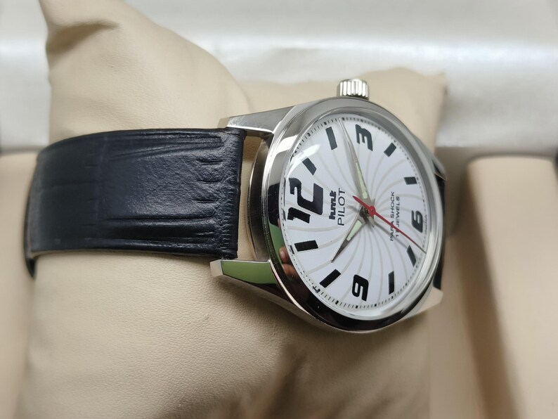 May include: A silver-toned wristwatch with a black leather band. The watch face is white with black numerals and hands, and the text "Pilot" is visible. The watch is displayed on a cream-colored surface.