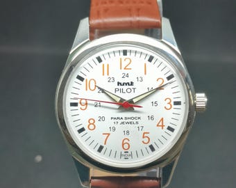 May include: A silver-toned wristwatch with a white face, featuring black and orange numerals and hands. The watch has a brown leather strap. The watch face includes the text "hmt PILOT" and "PARA SHOCK 17 JEWELS".