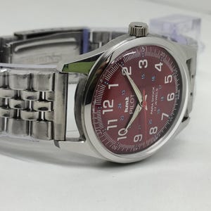 May include: A silver wristwatch with a burgundy dial. The watch face features white numerals, hands, and the text "hmt PILOT". The watch has a stainless steel band.