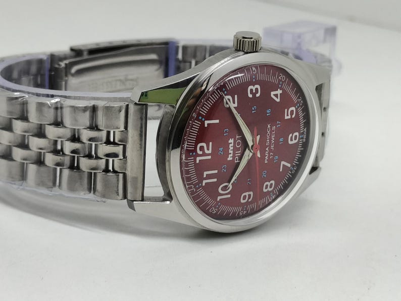 May include: A silver wristwatch with a burgundy dial. The watch face features white numerals, hands, and the text "hmt PILOT". The watch has a stainless steel band.