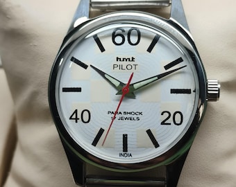 May include: A silver-toned wristwatch with a white face featuring black numerals and markers. The watch face displays the text "hmt PILOT" and "PARA SHOCK 17 JEWELS". The watch has a silver band and a red second hand.