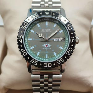 Könnte beinhalten: Eine Citizen Automatic Uhr mit einem silbernen Edelstahlarmband und einer schwarzen drehbaren Lünette. Das Zifferblatt ist grau mit Leuchtzeigern und -markierungen, und der Tag wird als "TUE 29" angezeigt. Die Uhr hat eine verschraubte Krone.