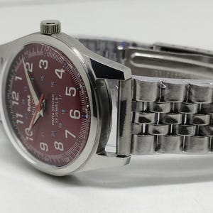 May include: A silver wristwatch with a burgundy dial featuring white numerals and hands. The watch has a stainless steel band and a crown at the 3 o'clock position. The watch face includes the text "PARA SHOCK 17 JEWELS".