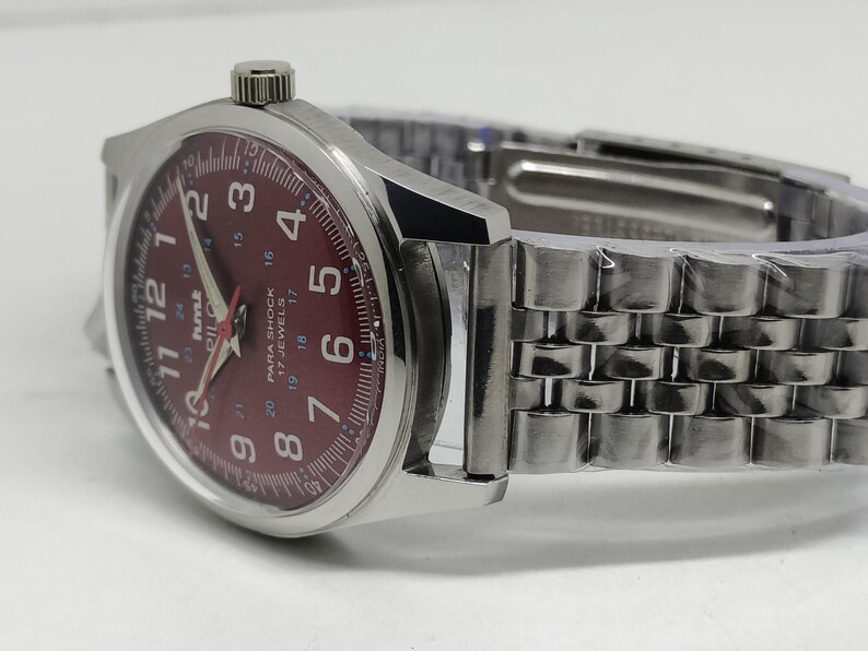 May include: A silver wristwatch with a burgundy dial featuring white numerals and hands. The watch has a stainless steel band and a crown at the 3 o'clock position. The watch face includes the text "PARA SHOCK 17 JEWELS".