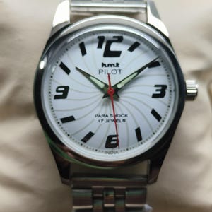 May include: Silver-toned wristwatch with a white face featuring black numerals and hands. The watch face displays the text "hmt PILOT" and "PARA SHOCK 17 JEWELS". The watch has a silver metal band.