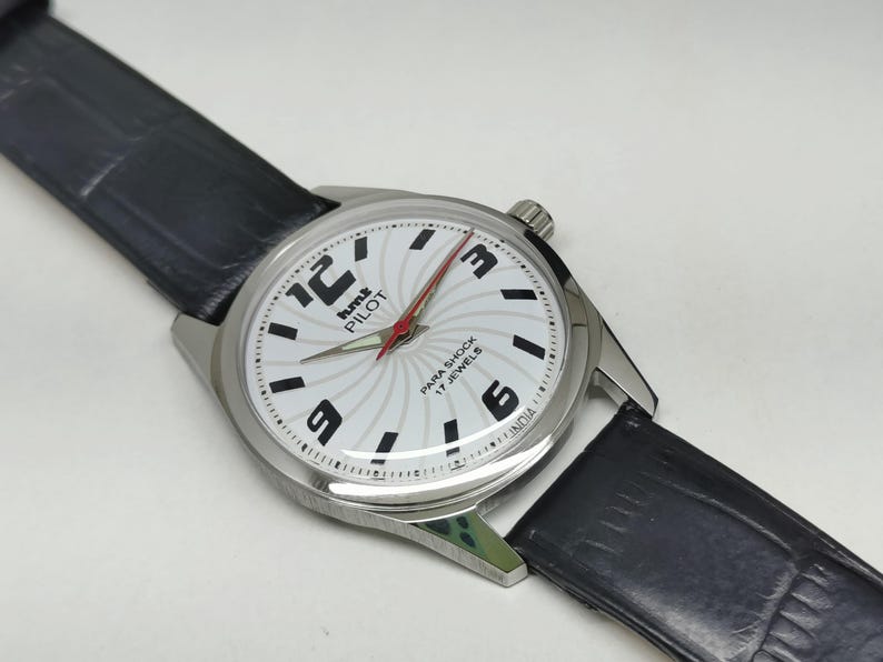 May include: A silver-toned wristwatch with a white face featuring black numerals and a red second hand. The watch has a black leather band. The watch face has the text "hmt PILOT PARA SHOCK 17 JEWELS INDIA".