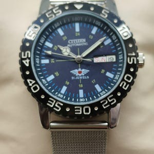 Può includere: Orologio da polso Citizen Automatic con quadrante blu scuro, numeri e lancette bianche. L'orologio ha una lunetta girevole nera e un cinturino in maglia d'argento. Il giorno è visualizzato come "DOM 19".